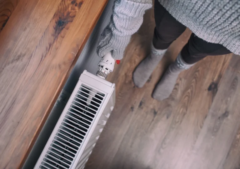 Person in warm clothing adjusting the temperature dial on a white radiator near a wooden surface, standing on a wooden floor.