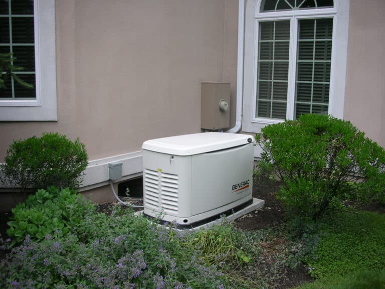 A standby generator is installed on a concrete pad outside a house, surrounded by bushes, near a window with white trim.