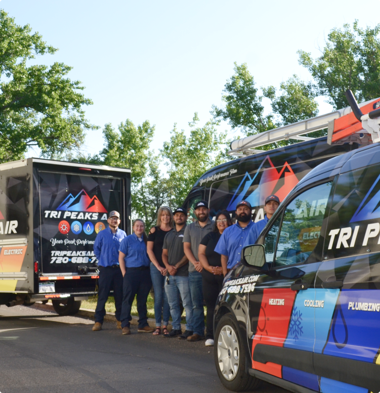 A group of people stand in front of Tri Peaks Air service vehicles parked on a road, with trees in the background.