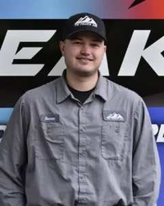 A man in a gray uniform and black cap stands in front of a vehicle with a company logo. The uniform has a name patch and a mountain logo on both the shirt and cap.