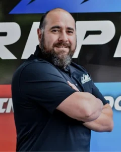 A man with a beard and mustache stands with arms crossed, wearing a dark polo shirt, in front of a colorful background with large text.