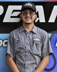 A smiling person wearing glasses, a black cap, and a gray button-up shirt stands in front of a multicolored sign with partial text.