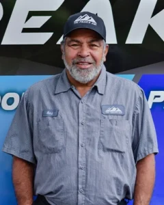 A man with a gray beard wearing a "Tri Peaks Air" cap and uniform shirt stands in front of a company-branded backdrop.