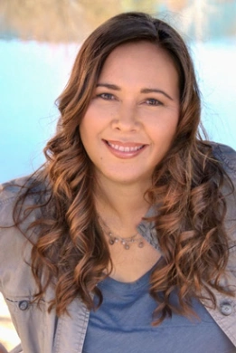 Woman with long wavy brown hair, wearing a blue top and gray jacket, smiling outdoors with a blurred natural background, enjoying the peace of mind that comes from a reliable maintenance plan.