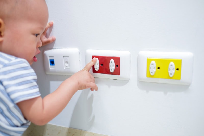 Electrical Safety Tips. Curious toddler baby boy child play with electric plug. Try to insert it into socket, Baby hand reach for electrical outlet covered with red safety plugs (child safety concept) - Selective focus.