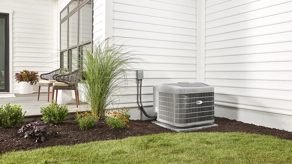 Carrier air conditioner at the side of a white house with landscaping and a patio in view.