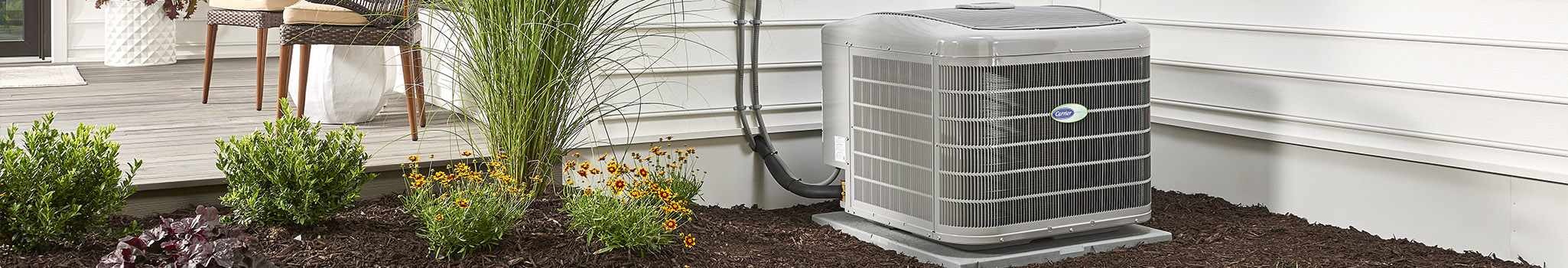 Carrier air conditioner at the side of a white house with landscaping and a patio in view.